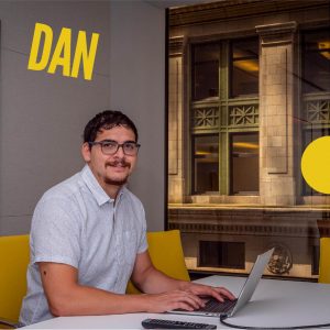 Man at computer in an office. Name Dan in yellow font.